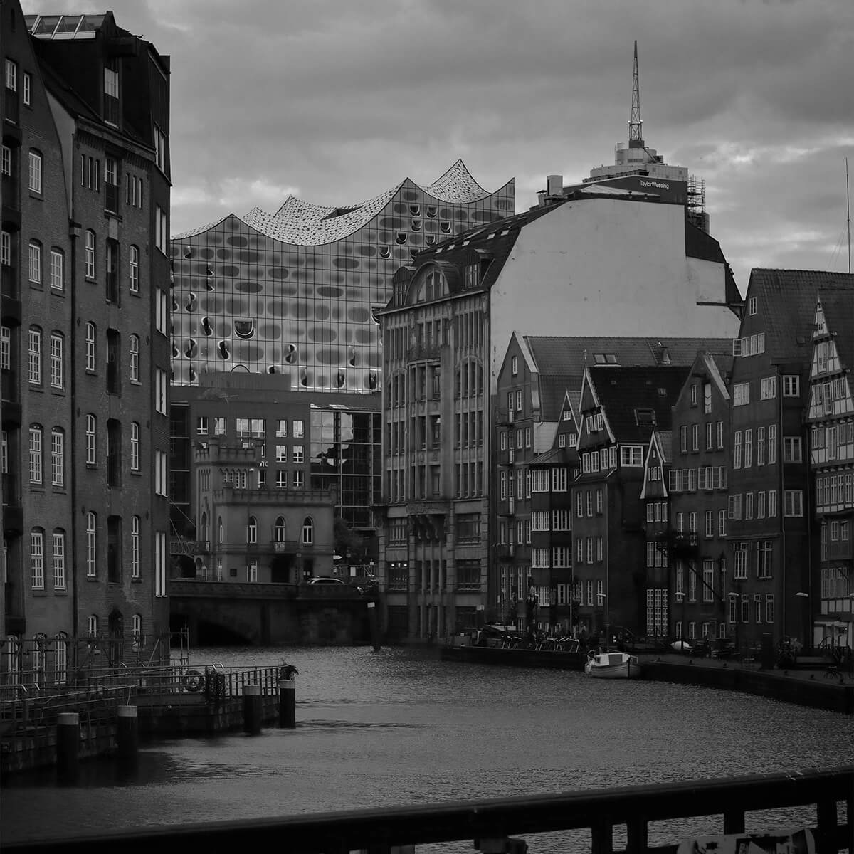 Schwarz-Weiß-Foto eines von historischen Gebäuden gesäumten Kanals, mit der modernen Elbphilharmonie mit Wellendach im Hintergrund unter einem bewölkten Himmel.