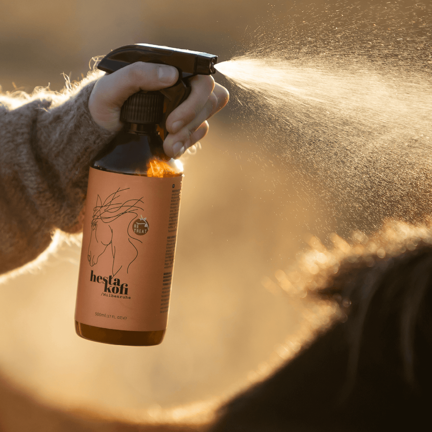 Eine Hand sprüht vor einem warmen, goldenen Hintergrund eine braune Flasche mit der Aufschrift Hestakofi Milbenruhe, auf der eine Strichzeichnung eines Pferdes abgebildet ist, auf das Fell eines Pferdes zur natürlichen Pflege durch Hestakofi.