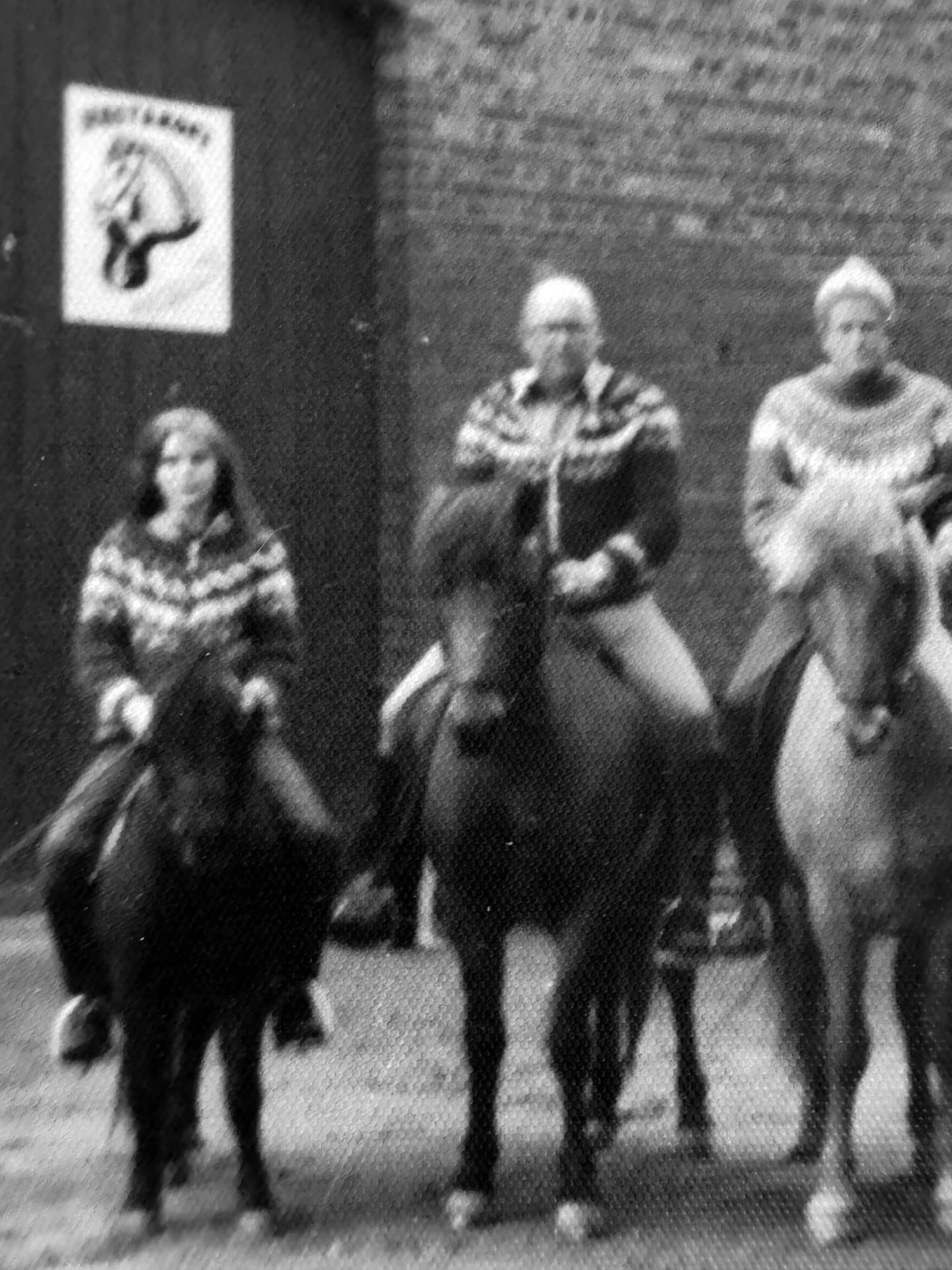 Drei Personen in gemusterten Pullovern reiten nebeneinander auf Pferden vor einem Backsteingebäude mit einem Schild, auf dem ein Pferdekopf abgebildet ist. Die Szene scheint schwarz-weiß oder in Graustufen zu sein.