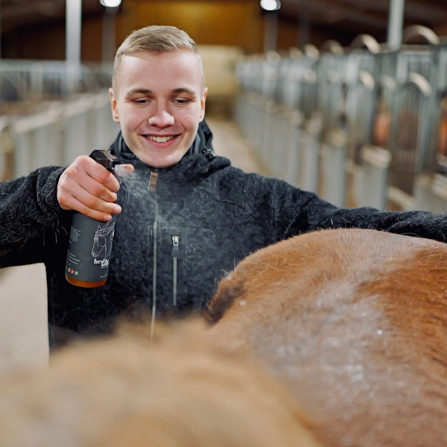 Ein Mann lächelt, während er in einem Stall Hestakofi Flake Away auf den Rücken eines Pferdes sprüht. Das Produkt fördert die Feuchtigkeit und unterstützt die Hautschutzbarriere des Pferdes. Im Hintergrund sind Ställe und sanfte Beleuchtung zu sehen.