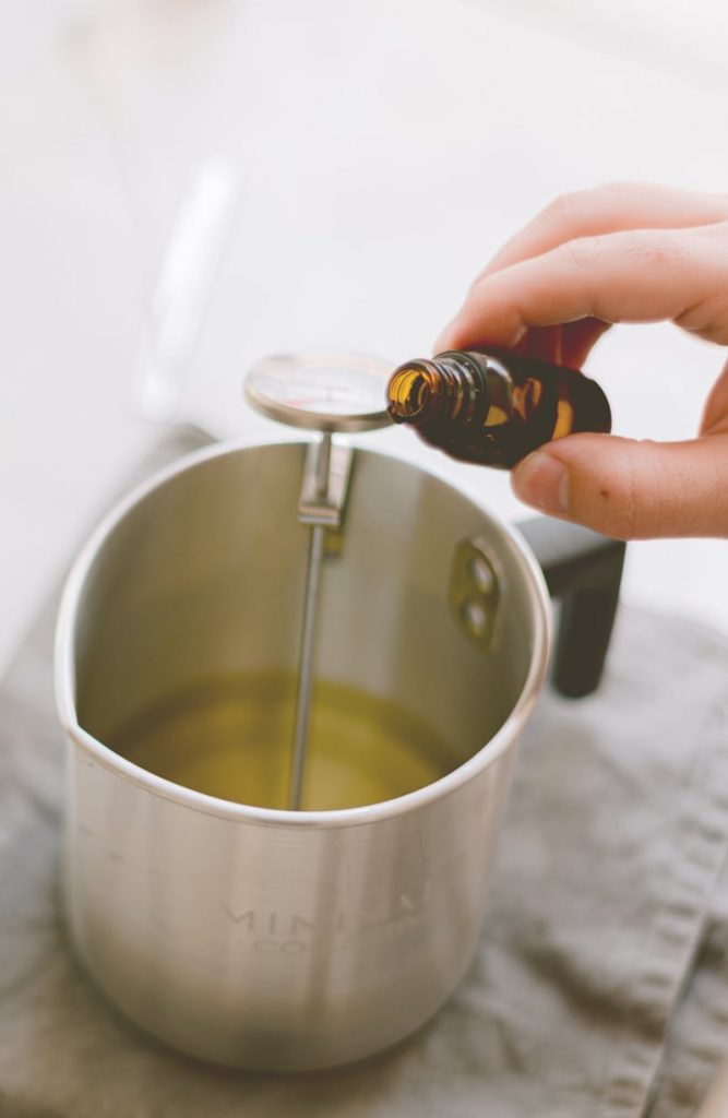 Eine Hand hält eine kleine braune Flasche und gießt Flüssigkeit in einen Topf aus rostfreiem Stahl, in dem sich ein Thermometer befindet. Der Topf enthält eine gelbliche Flüssigkeit und ruht auf einem grauen Tuch.