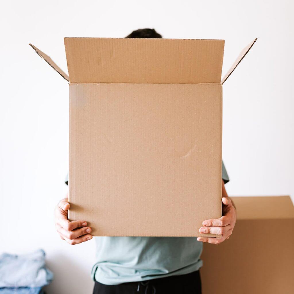 Eine Person hält einen großen offenen Karton vor ihr Gesicht, der ihre Identität verdeckt, vor einem schlichten weißen Hintergrund. Im Hintergrund sind ein weiterer Karton und einige gefaltete Kleidungsstücke zu sehen.