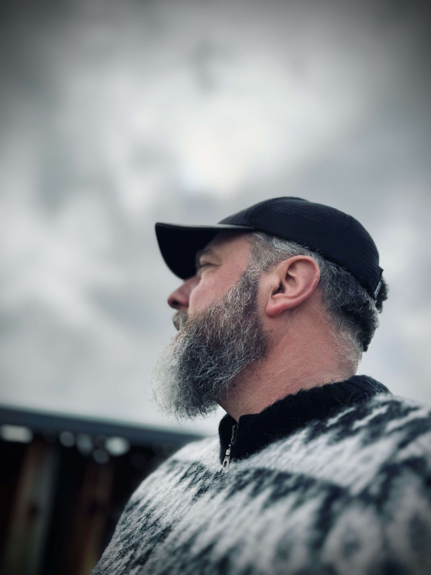 Ein bärtiger Mann, der eine schwarze Mütze und einen gemusterten Pullover trägt, blickt vor einem wolkigen, unscharfen Himmelshintergrund zur Seite. Das Bild ist sanft fokussiert und schafft eine stimmungsvolle Atmosphäre.