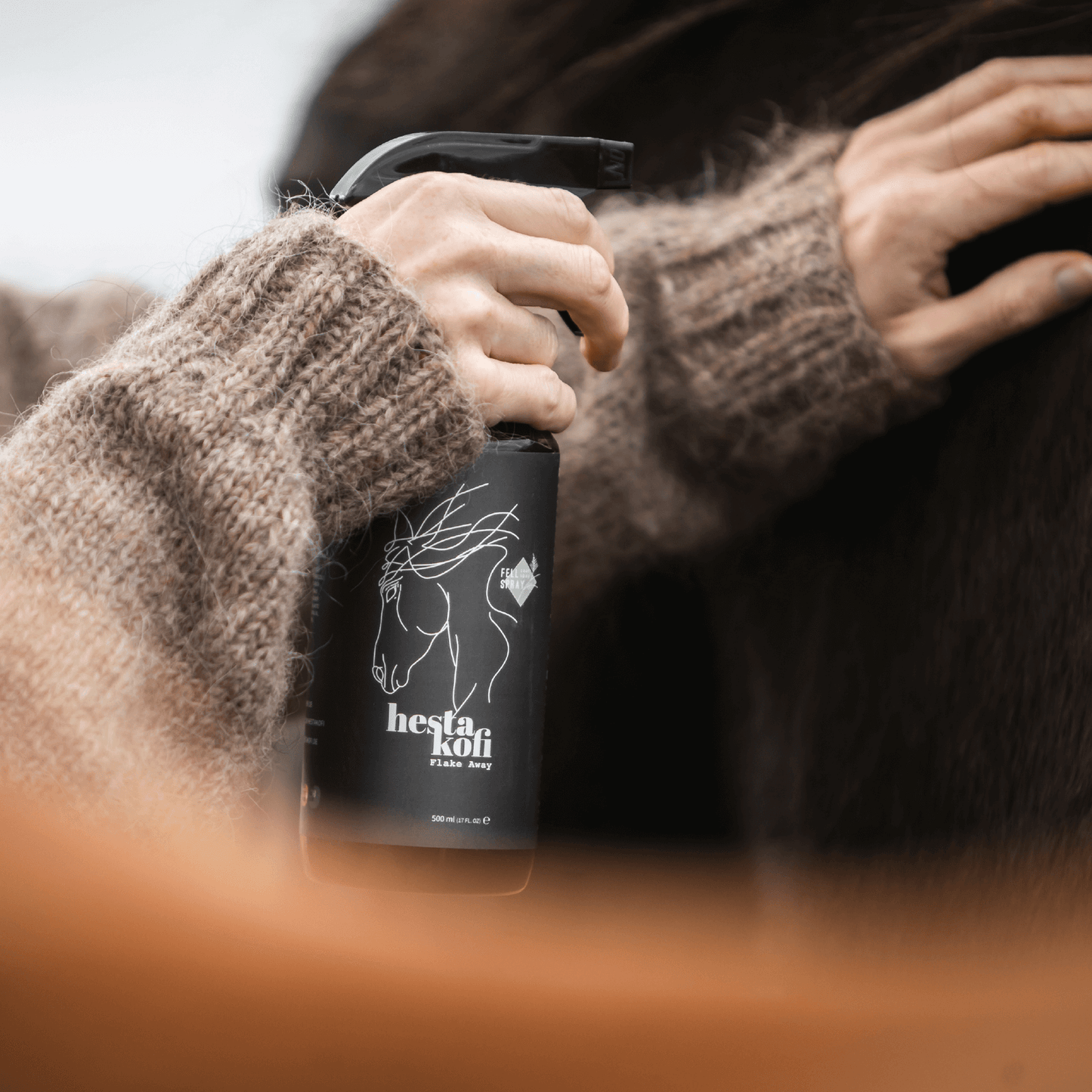 Eine Person in einem braunen Wollpullover sprüht Hestakofi Flake Away, eine schwarze Flasche mit einer Pferdeabbildung, auf die Seite eines dunkelbraunen Pferdes, um eine optimale Hydratation und Glanz zu fördern.