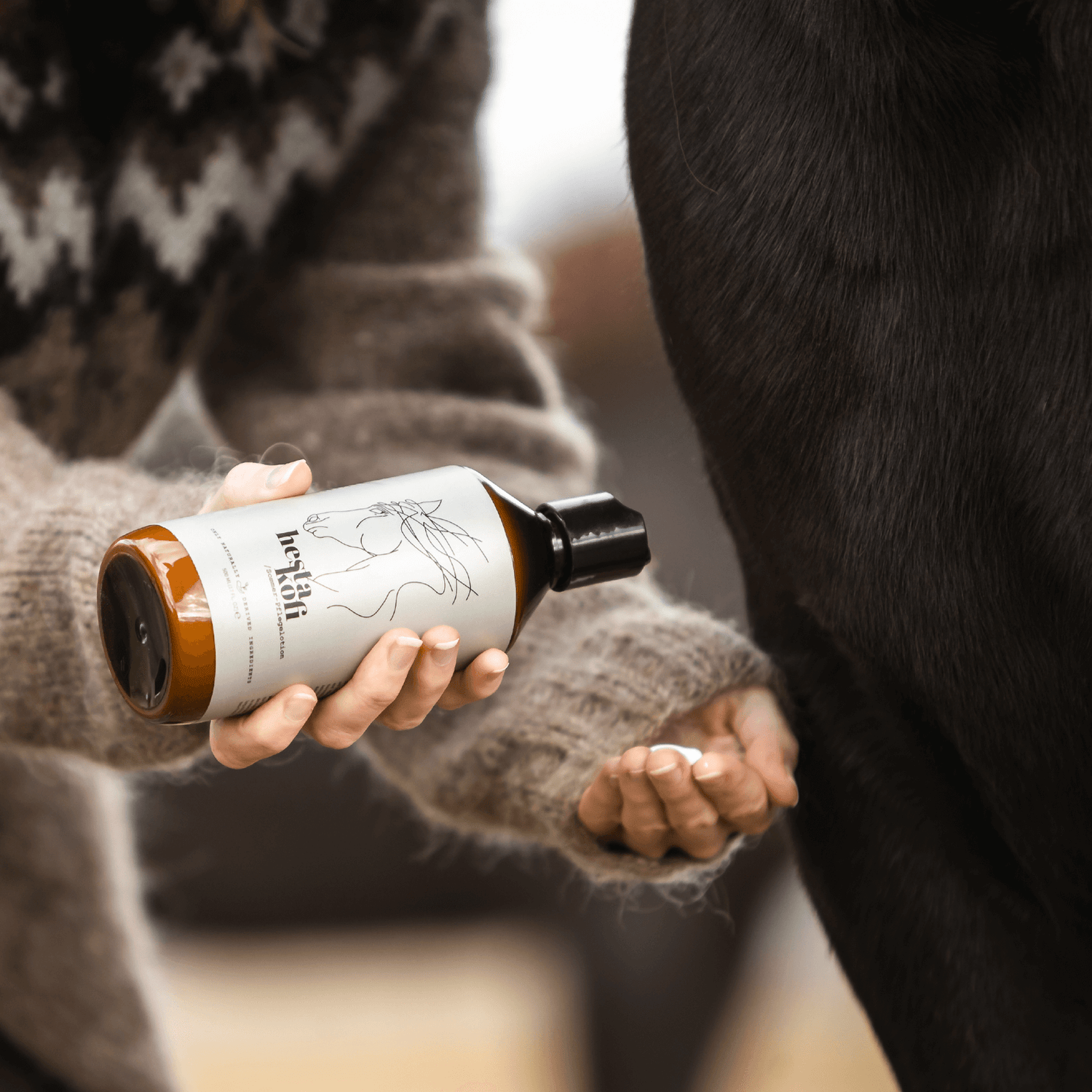 Eine Person in einem Island Wollpullover dosiert Hestakofi Sommerpflegelotion, eine braune Flasche mit einer Pferdeabbildung, in die Hand wir sehen die Lweisse Lotion